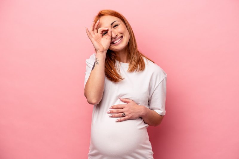 mulher gravida em fundo rosa fazendo o símbolo de "ok" na frente do rosto
